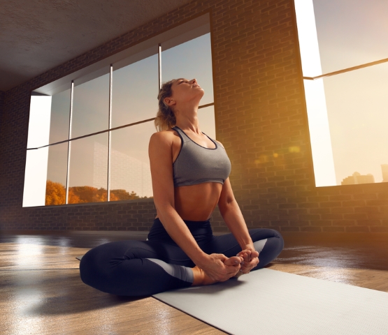 Femme pratiquant le yoga en intérieur sur un tapis, assise en posture de relaxation dans une salle lumineuse avec de grandes fenêtres