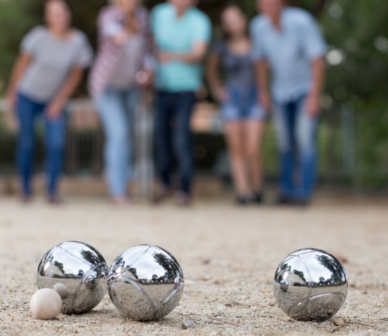 Boules de pétanque sur un terrain en gravier au premier plan, avec un groupe de personnes en arrière-plan flou en train de jouer
