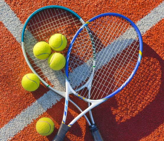 Deux raquettes de tennis croisées formant un cœur avec plusieurs balles de tennis posées sur un court en surface rouge