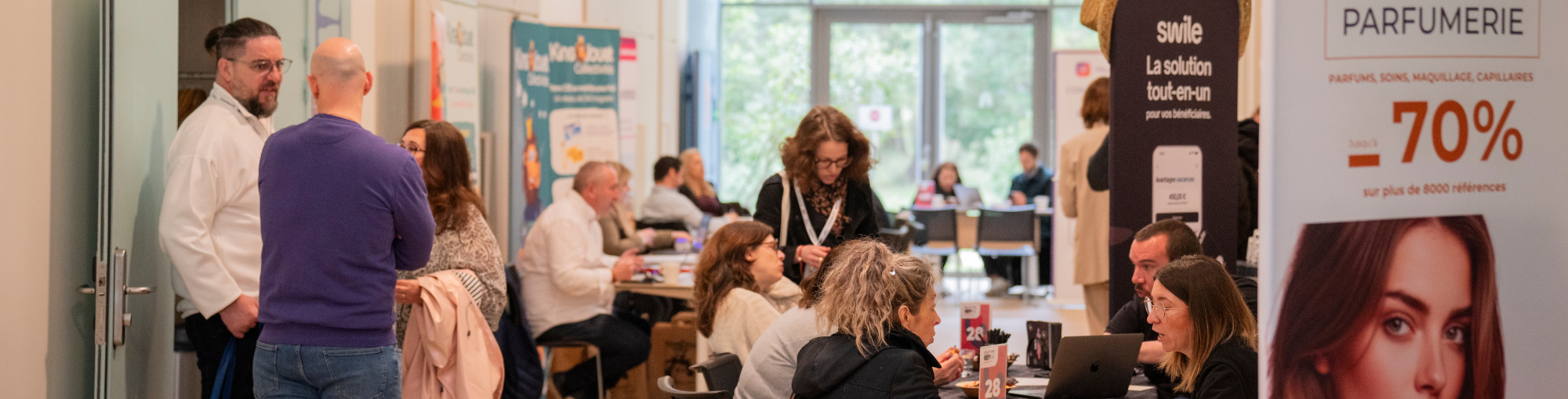 Une scène de salon professionnel ou de networking : plusieurs personnes discutent autour de stands promotionnels dans un espace lumineux et convivial.