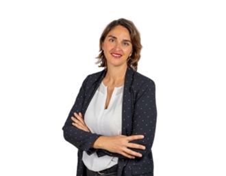 Portrait professionnel de Delphine Joly, femme souriante aux cheveux courts, portant une veste sombre à pois et un chemisier clair, bras croisés sur fond neutre.